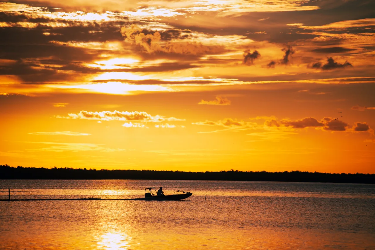 A silhouetted boat crossing glowing water beneath an amber sunset